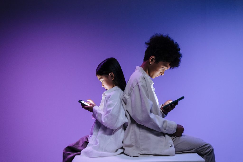 Duas crianças usam smartphones lado a lado sobre um fundo roxo.