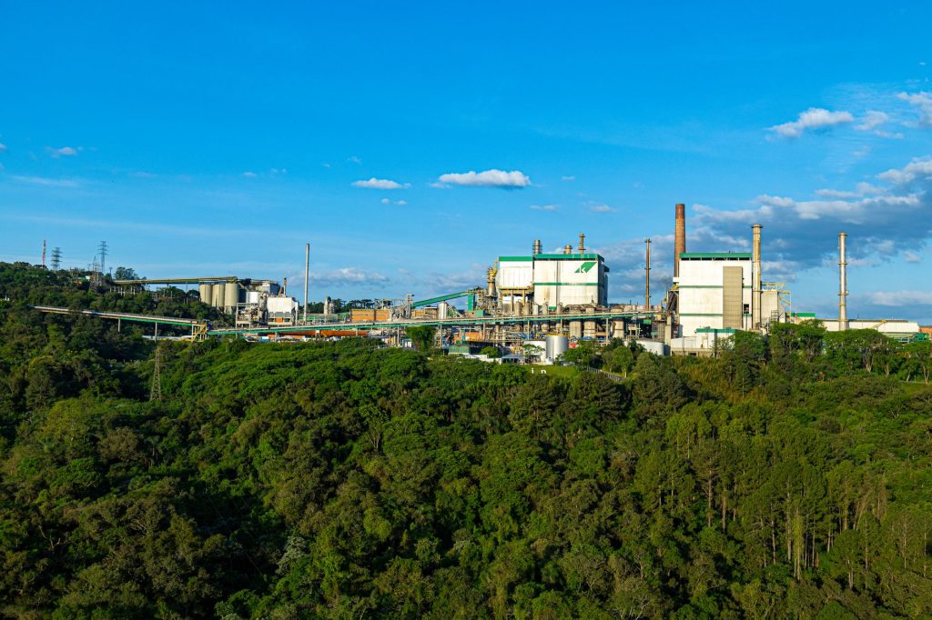 Vista aérea ampla de uma planta industrial cercada por área de floresta em Telêmaco Borba, no Brasil.